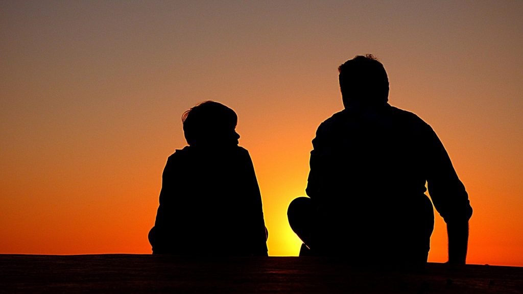 a father and son sit, watching a sunset